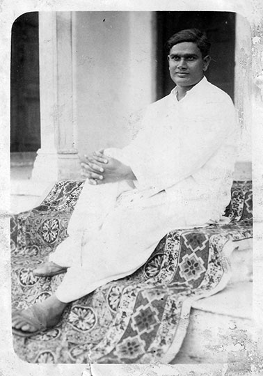 A black and white photograph of Babu as a young man. He is dressed all in white and is seated on stairs that are covered with a traditional rug.