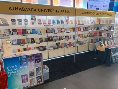 An exhibition booth for Athabasca University Press featuring shelves filled with books and a banner highlighting new and forthcoming titles.