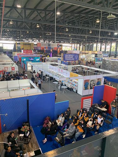 A high-angle view of a large, busy book fair in Mexico City, showing various publisher booths and an audience seated for a presentation.